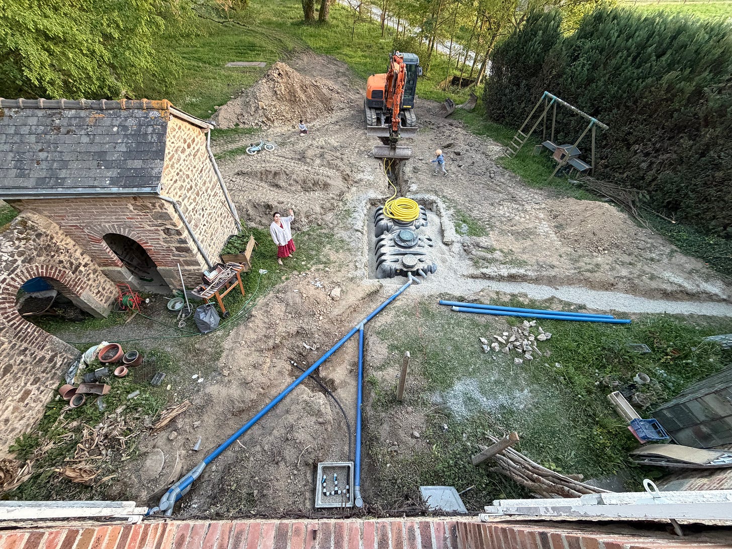 Aerial view of a garden torn up for a septic installation: an orange digger at the end of a long trench, a large black tank set in gravel with a coil of yellow pipe on top, blue pipes running across the dirt, a stone outbuilding with brick arches on the left, a woman in a red skirt waving up at the camera, and a wooden swing set at the edge. Aerial view of a garden torn up for a septic installation: an orange digger at the end of a long trench, a large black tank set in gravel with a coil of yellow pipe on top, blue pipes running across the dirt, a stone outbuilding with brick arches on the left, a woman in a red skirt waving up at the camera, and a wooden swing set at the edge.