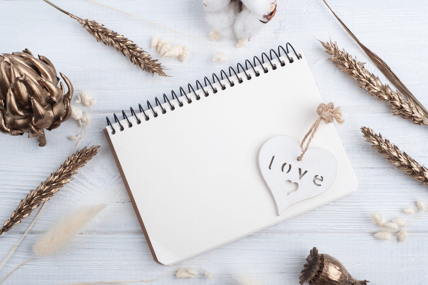 Blank notebook over white wooden background, surrounded by golden flowers. Over the notebook there’s a white wooden heart with the word “love” and a small heart carved out.