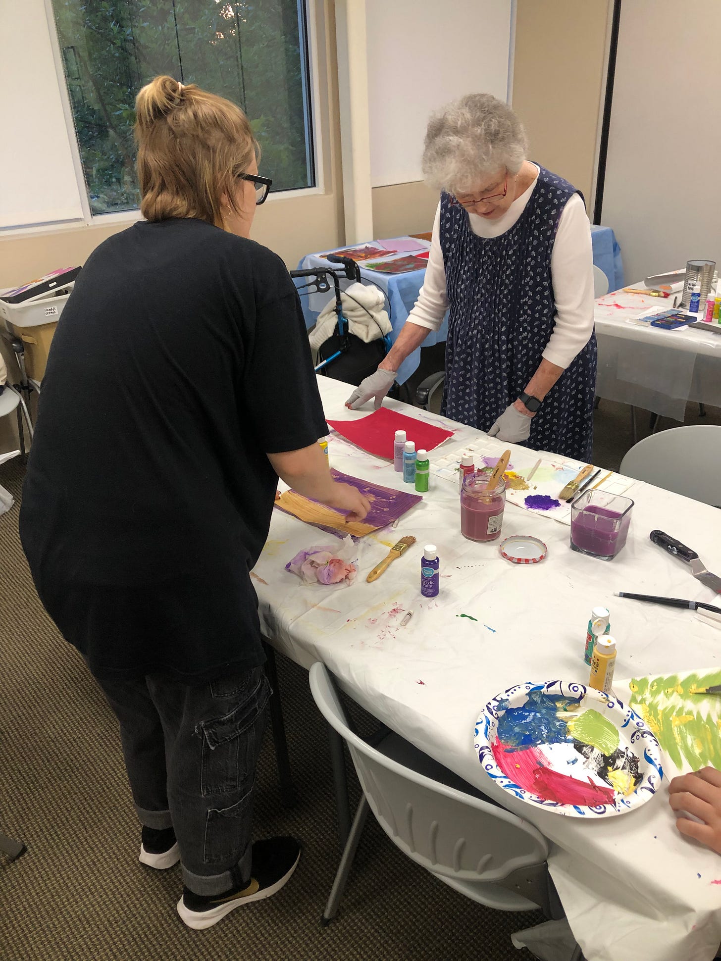 Shady wearing a black outfit is talking to her grandmother who is wearing a blue dress and gloves to protect her hands. On the table are various painting supplies.