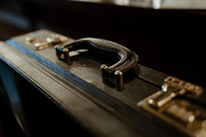 A close-up of the top of an old-style, hard-sided leather briefcase, showcasing the polished brown handle and the brass locking latch hardware. The background is dark and unclear, making the case itself seem more like a symbol of a certain kind of life than like an actual object taking up actual space. A close-up of the top of an old-style, hard-sided leather briefcase, showcasing the polished brown handle and the brass locking latch hardware. The background is dark and unclear, making the case itself seem more like a symbol of a certain kind of life than like an actual object taking up actual space.