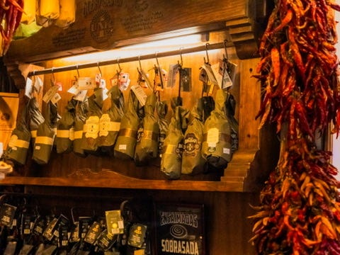 “Close-up of a wooden display with sobrasada sausages wrapped in paper, hanging alongside strings of dried red peppers.”