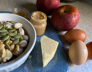 bowl of nuts with cheese next to peanut butter, apples, and eggs