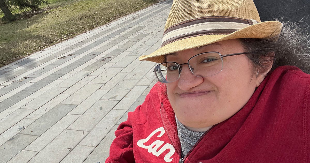 Athena smiling closed-mouth while sitting in her wheelchair on an outdoor pathway. She's wearing a red Canada hoodie and a wicker fedora-style hat.