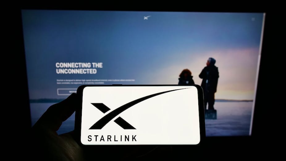 A hand holding a smartphone that features a black x and the word Starlink against a white background in front of a larger screen that reads Connecting the unconnected A hand holding a smartphone that features a black x and the word Starlink against a white background in front of a larger screen that reads Connecting the unconnected