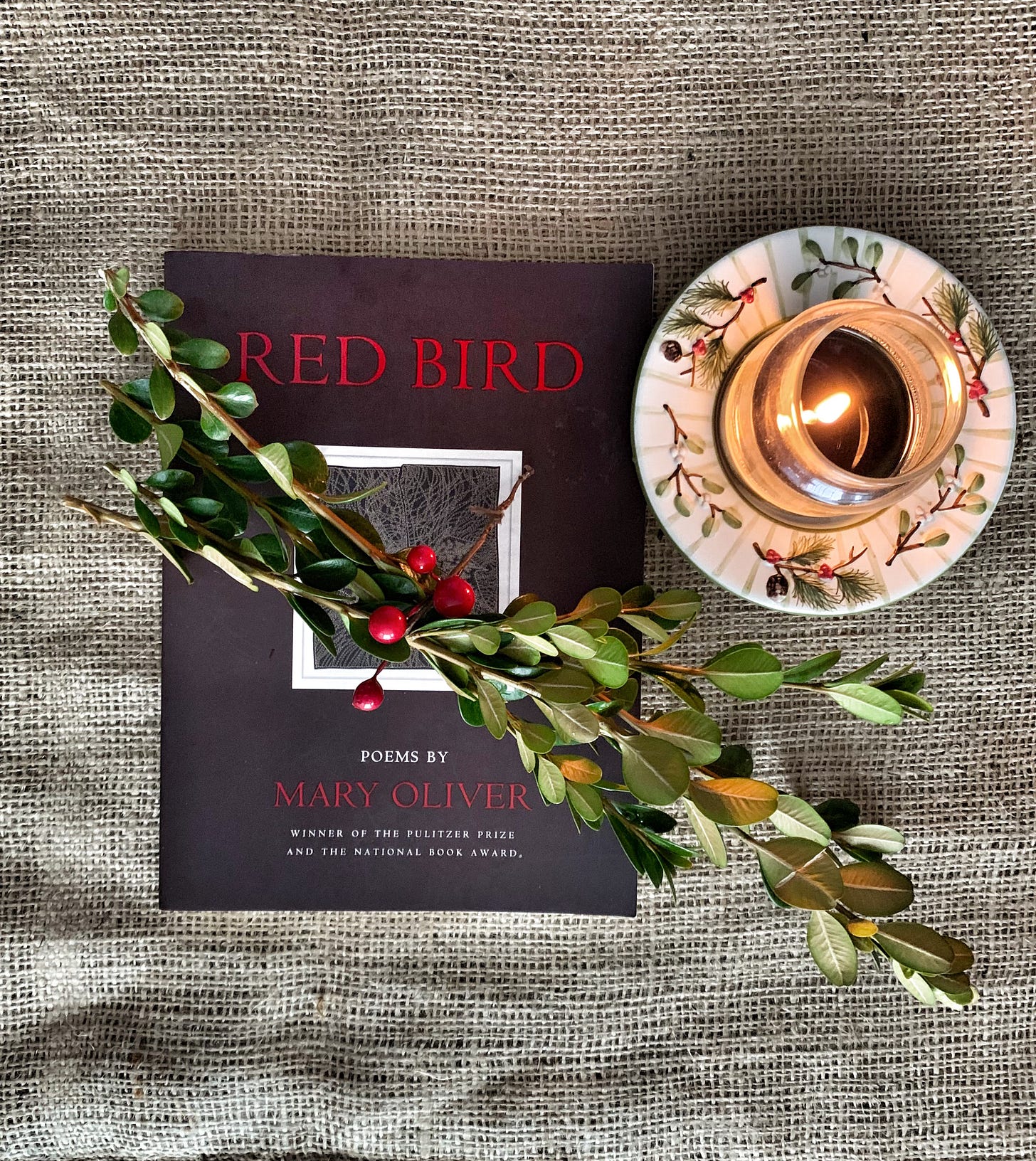 A copy of Mary Oliver's book of poems "Red Bird" on a muslin cloth beside a lit candle and a bundle of boxwood stems 