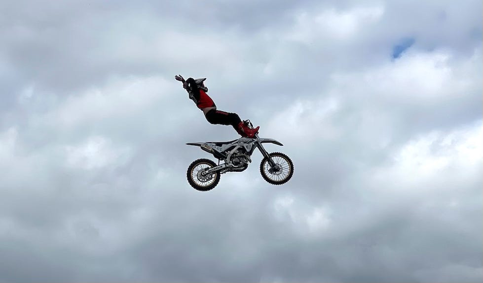 A motocross rider in mid-air performs a daring stunt, fully extended off the back of their dirt bike with arms reaching upward, while the motorcycle soars against a cloudy sky.