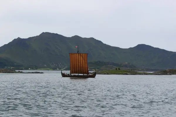 A Viking-Age wooden ship sails in the Lofoten Islands, Norway. It has a yellow and red sail and is pictured in front of mountains and a grey sky