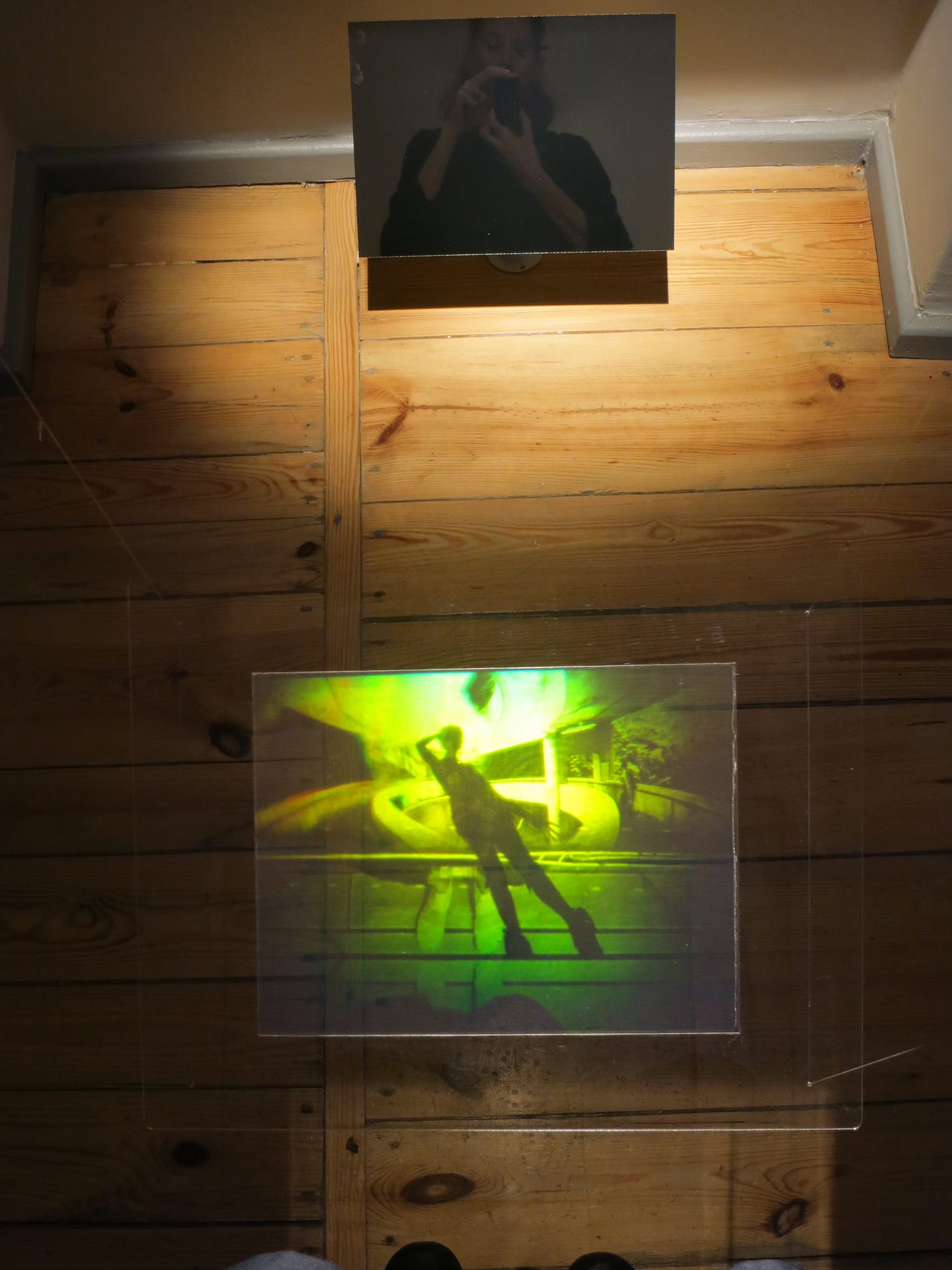 In the lower part of the photograph is a transmission hologram, with light shining through it to created an image of green and yellow hues. The hologram is a montaged image that includes the feet and shadow of the photographer. In this photograph of the hologram we can also see the feet and the reflection of the photographer (who in both cases is the artist Martina Mrongovius