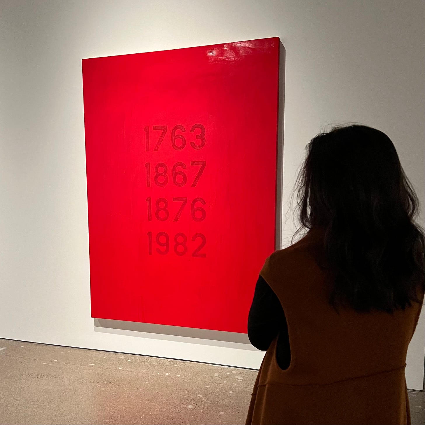 The back of a woman standing to the right side and in front of a painting by Robert Houle. The painting is red with four years on it. The back of a woman standing to the right side and in front of a painting by Robert Houle. The painting is red with four years on it.