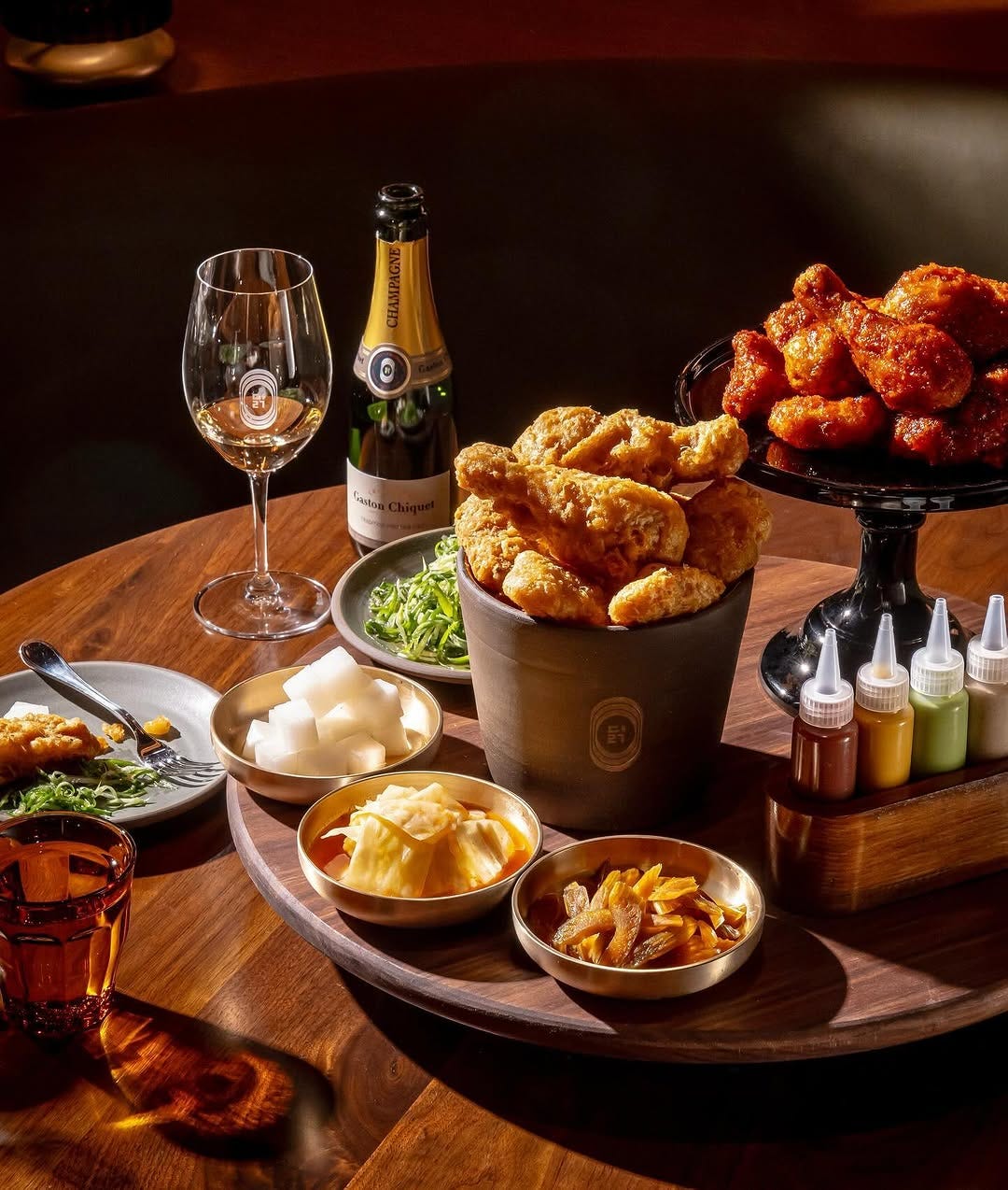 A table with two types of korean fried chicken, a bottle of champagne, four different colored sauces in squeeze bottles, and assorted banchan A table with two types of korean fried chicken, a bottle of champagne, four different colored sauces in squeeze bottles, and assorted banchan