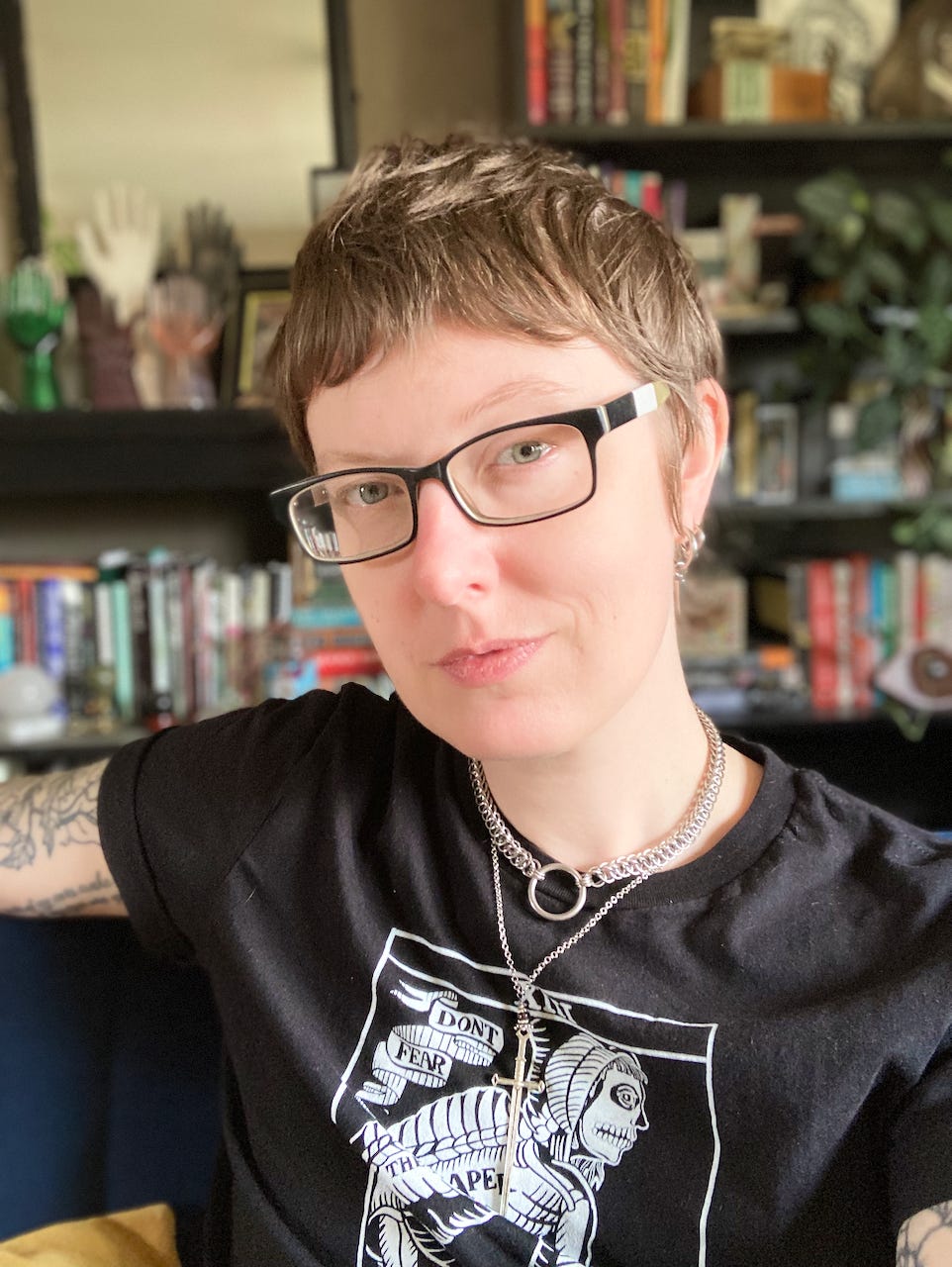 Charlie, a white androgynous person with dark blond hair, green eyes, and arm tattoos, sits on a blue couch with book and plant-filled bookshelves behind them. They are smiling faintly and wearing a black t-shirt.