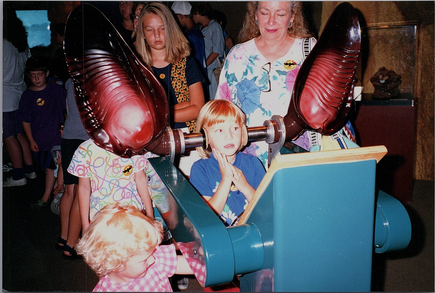 A small child has her head between two giant bat ears at a museum exhibit. There is a line of people behind her waiting to try them on.