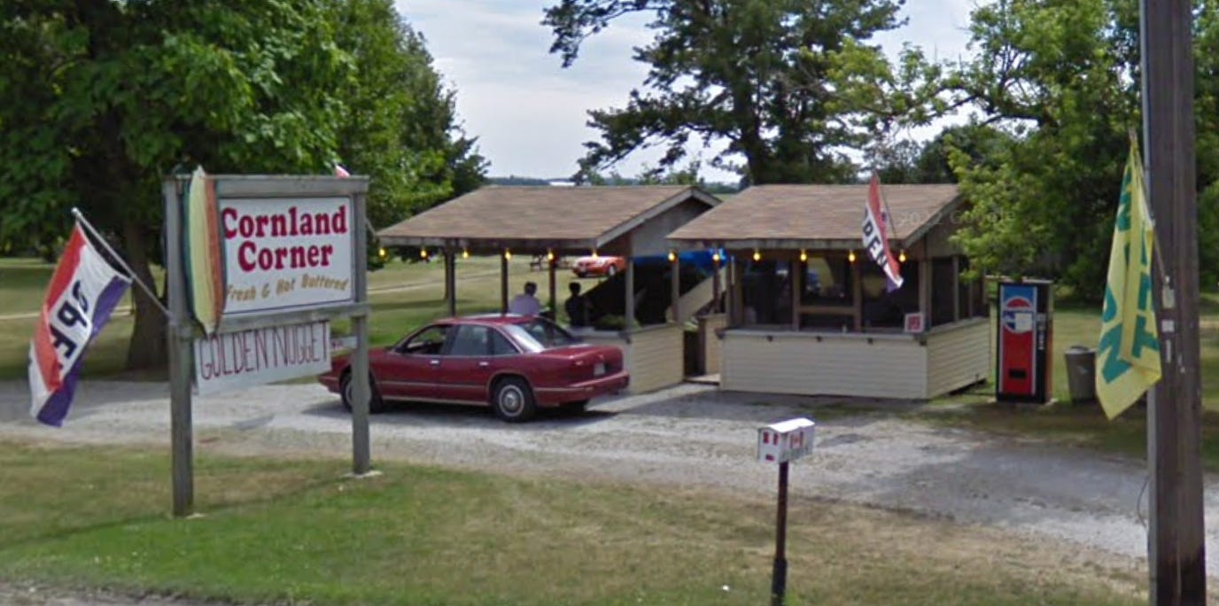 A red car is parked in front of two small, yellow corn stands. There's a Pepsi vending machine beside them. In the foreground there is a roadside sign that reads, "Cornland Corner - Fresh and Hot Buttered" There is an image of an ear of corn on the sign and a flag that says "OPEN" is attached to it as well. A red car is parked in front of two small, yellow corn stands. There's a Pepsi vending machine beside them. In the foreground there is a roadside sign that reads, "Cornland Corner - Fresh and Hot Buttered" There is an image of an ear of corn on the sign and a flag that says "OPEN" is attached to it as well.