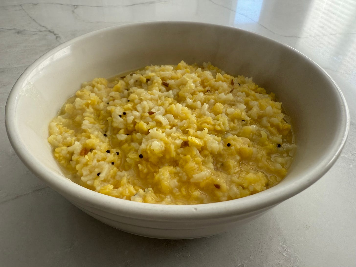A warm bowl of kitchari—rice and mung dal with mustard seeds and cumin—in a white ceramic bowl on a marble surface, with soft natural light.