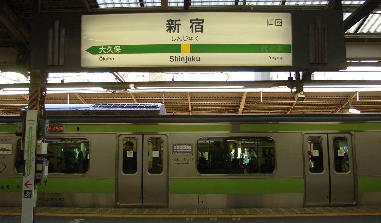 A green-liveried JR Yamanote Line train stopped at Shinjuku station platform, with a large overhead station sign in Japanese and English reading 'Shinjuku,' showing Ōkubo as the previous stop and Yoyogi as the next, and passengers visible through the train windows. A green-liveried JR Yamanote Line train stopped at Shinjuku station platform, with a large overhead station sign in Japanese and English reading 'Shinjuku,' showing Ōkubo as the previous stop and Yoyogi as the next, and passengers visible through the train windows.