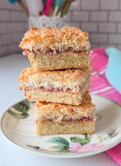 a pink baking tray with almond macaroons and glace cherries on it. A cooling rack with apple and blackberry muffins on it with apples at the top right hand of the shot. A pink cake plate with a 4 layer spiced plum upside down cake on it.