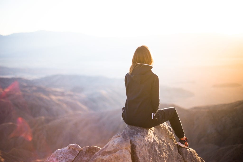 person sitting on top of gray rock overlooking mountain during daytime person sitting on top of gray rock overlooking mountain during daytime