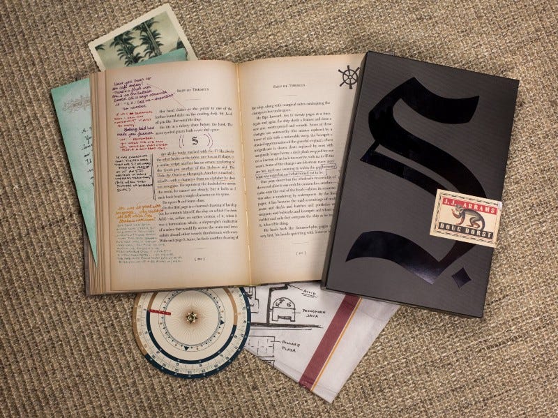 Descrição da imagem: a foto mostra o conjunto da publicação. Existe um livro aberto, com páginas amareladas e anotações à caneta feita nas margens, guardanapos desenhados, uma bússola, um pedaço de uma carta, um pedaço de uma foto e uma capa, preta com o símbolo de um S. e um macaco, onde o livro é guardado.