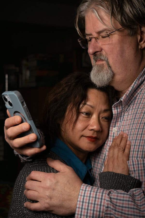 An East Asian woman sits on a couch next to a white man, leaning her head on his cheek.