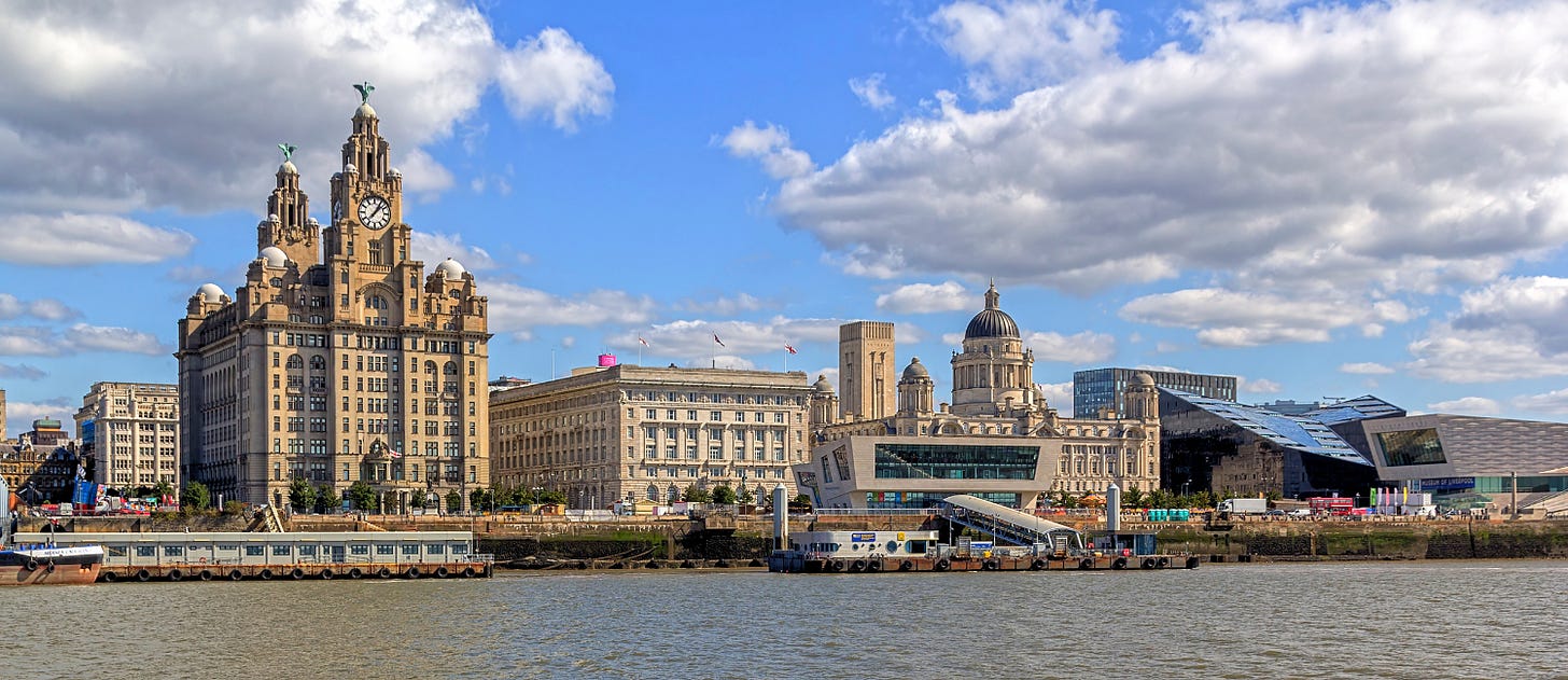 Panoramic view of Liverpool’s waterfront along the River Mersey, featuring the iconic Three Graces — the Royal Liver Building with its twin clock towers topped by Liver Birds, the Cunard Building, and the Port of Liverpool Building. Modern structures like the Museum of Liverpool and the glass-fronted Mann Island buildings contrast with the historic architecture under a bright blue sky with scattered clouds.