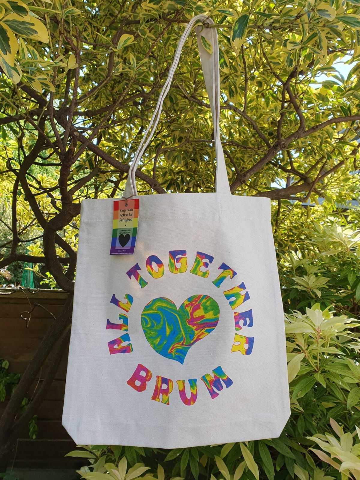 A tote bag hanging from a tree - the bag features a heart and the text All Together Brum, and is tie-die vibrant colours.