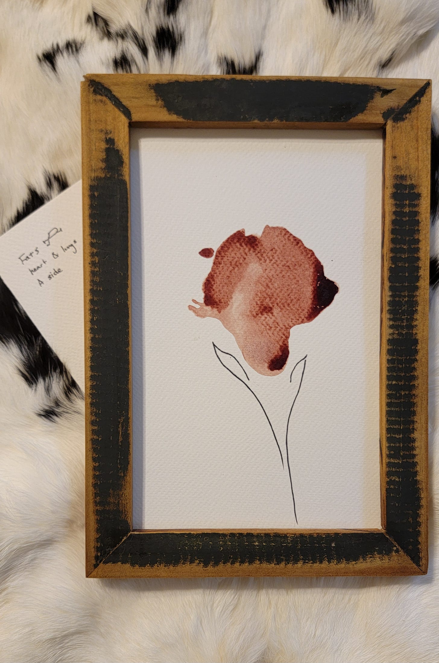 A red floral stain on white paper, framed in a distressed black wooden frame. All sitting against a black-spotted, white rabbit pelt.