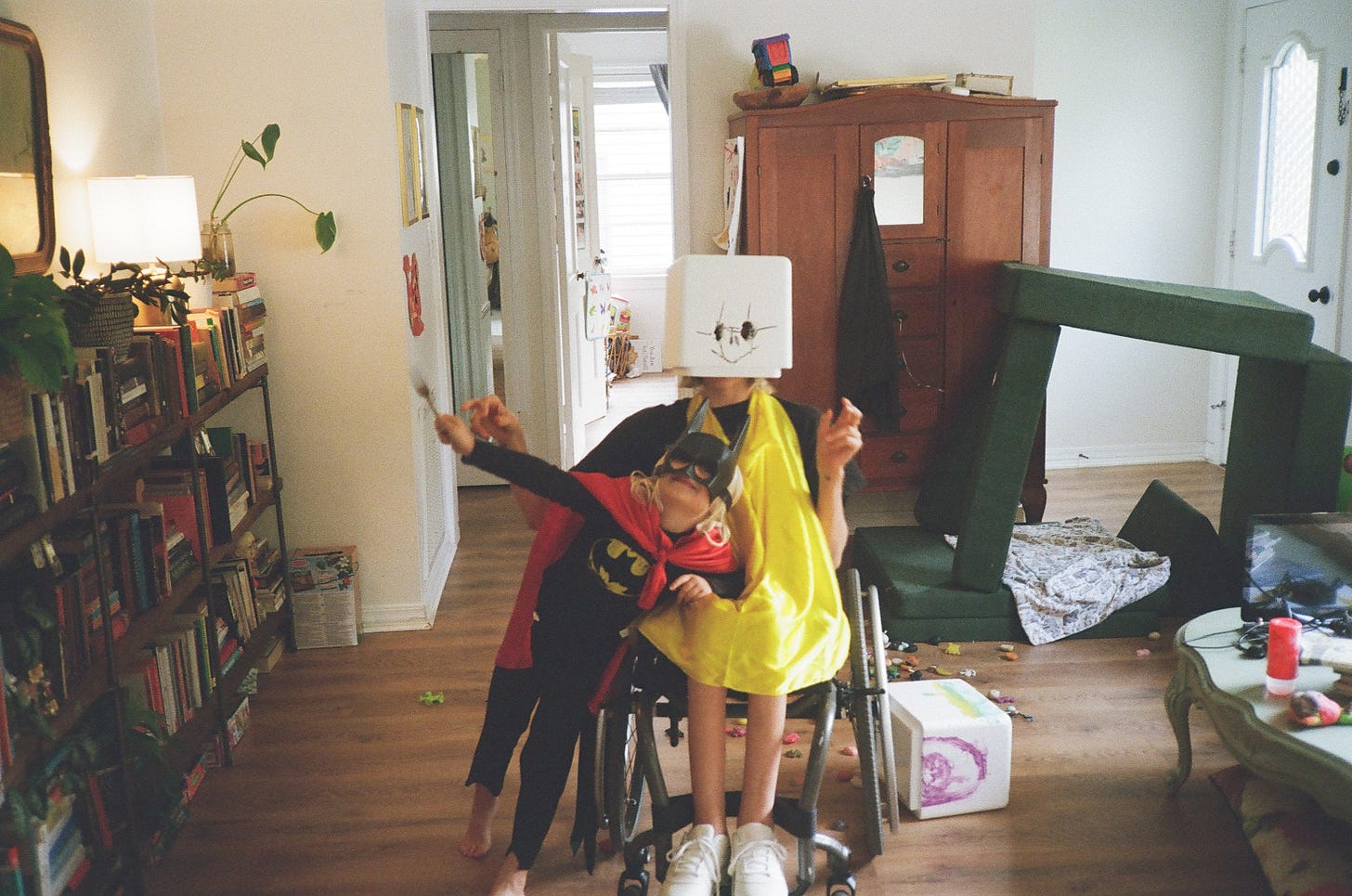Rebekah sits in her wheelchair, a yellow cape hanging from her neck, a homemade mask over her head. Her son leans against her lap wearing a batman costume and mask, a red cape draped across his back, a blurry fork in his hand. There is a bit of a chaos mess around them. Rebekah sits in her wheelchair, a yellow cape hanging from her neck, a homemade mask over her head. Her son leans against her lap wearing a batman costume and mask, a red cape draped across his back, a blurry fork in his hand. There is a bit of a chaos mess around them.