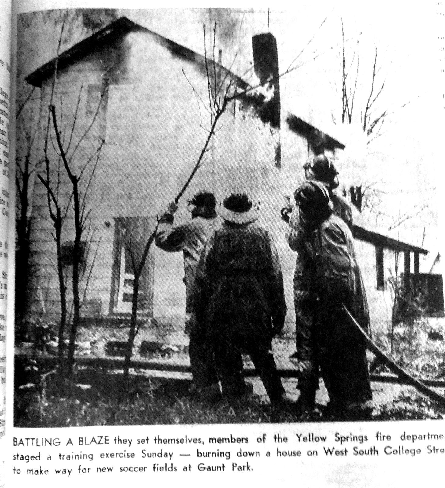 Newspaper photo and caption. Photo shows four firefighters in helmets, one holding a hose, looking at a two-story house in flames. The caption reads: BATTLING A BLAZE they set themselves, members of the Yellow Springs fire department staged a training exercise Sunday burning down a house on West South College Stre to make way for new soccer fields at Gaunt Park.