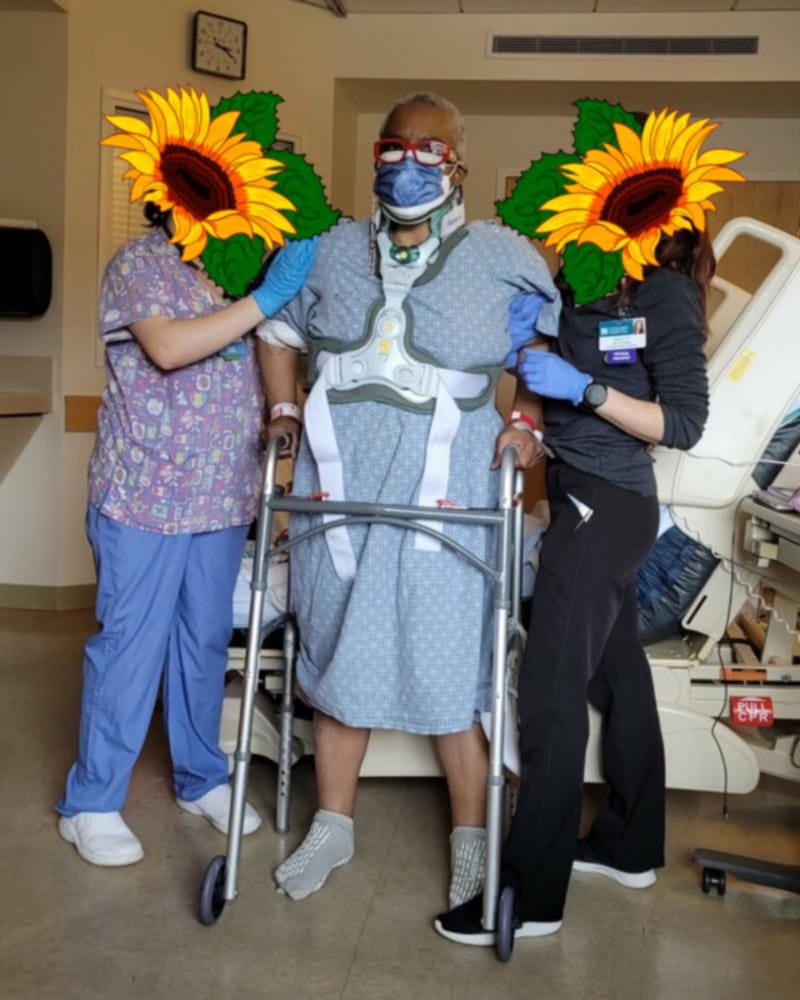 A photo of Claire in a hospital gown. Over the hospital gown is a neck and torso brace, akin to a car crash brace. She’s also wearing a surgical mask over an N95, and her signature red glasses. Two nurses, covered with sunflower illustrations, are holding her up and she stands in front of a hospital bed. Claire is gripping onto a walker. A photo of Claire in a hospital gown. Over the hospital gown is a neck and torso brace, akin to a car crash brace. She’s also wearing a surgical mask over an N95, and her signature red glasses. Two nurses, covered with sunflower illustrations, are holding her up and she stands in front of a hospital bed. Claire is gripping onto a walker.