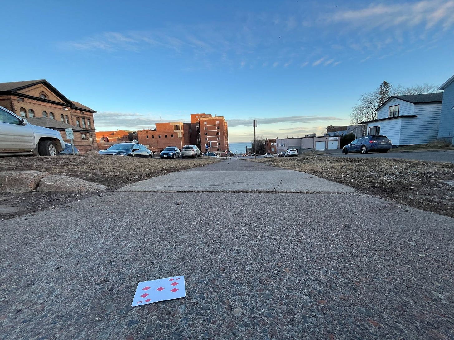 The sidewalks of Duluth, a lone playing card lays face up: the five of diamonds. The blue sky stands out against the muted grey that dominates the ground, the lake peers out in a small opening between the buildings.