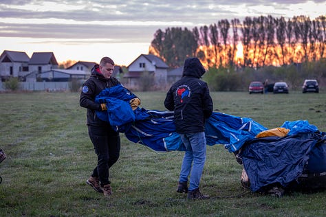 Înainte ca baloanele să urce, totul începe jos, pe iarbă, în frigul subțire al dimineții sau în lumina care abia se stinge peste câmp. Oamenii își ocupă locurile, verifică arzătoarele, întind pânza, ridică nacela, privesc cerul și, mai ales, ascultă vântul. În jur, mașinile stau cu farurile aprinse.