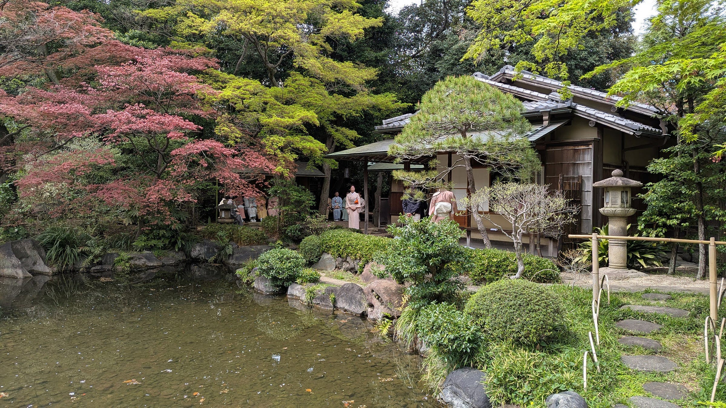 Lush trees and a stone lantern surround a Japanese tea house facing a pond