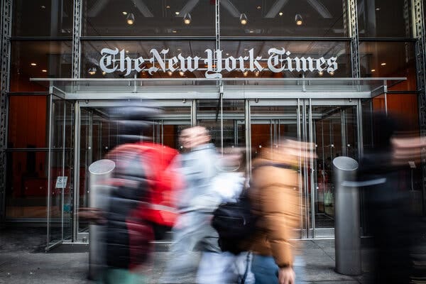 People walking in front of a glass building with the words “The New York Times” above the doors.