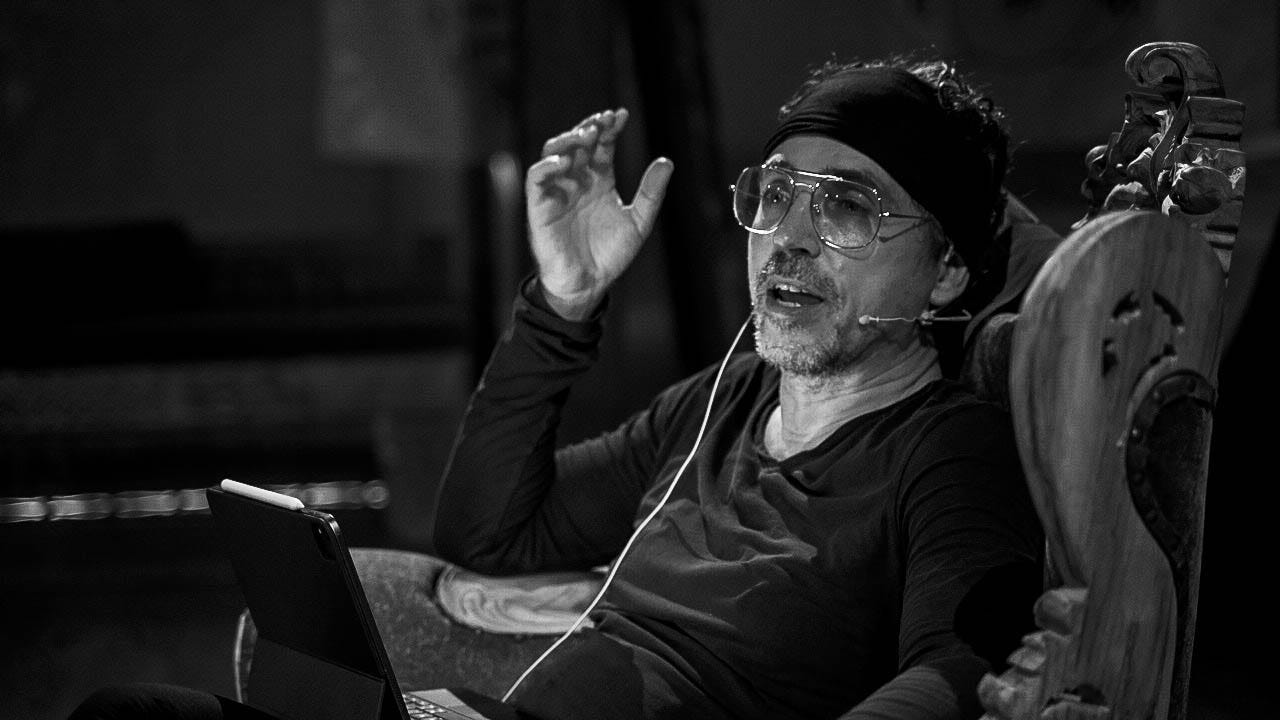 Black and white photograph of Michael Perin Wogenburg sitting on an ornate chair, wearing glasses and a headband, gesturing with his hand while speaking into a headset microphone during a nighttime talk on ritual color practice