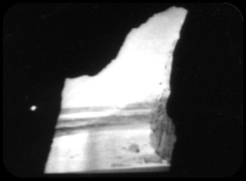 A monochrome photograph taken from a television screen showing the view from inside a cave, the entrance of which frames the image, looking out onto a beach and the sea in the distance.