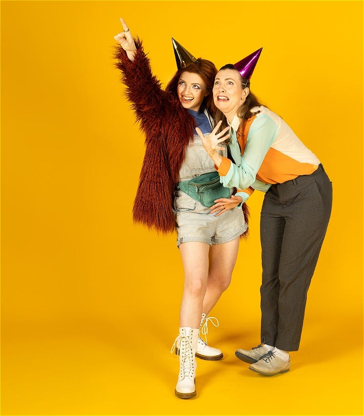 Two women on a bright yellow background, they both wear party hats and point at the sky out of frame.