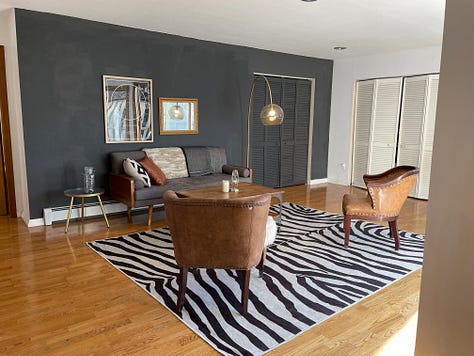each photo is of Andrea and Meg's living room, painted in progressively different colors with slight alterations on decor at well. In first photo, wall is charcoal colored. Subsequently, emerald, light pink, wallpapered, white with a "stay tender sign", then white with a triptych of cacti.