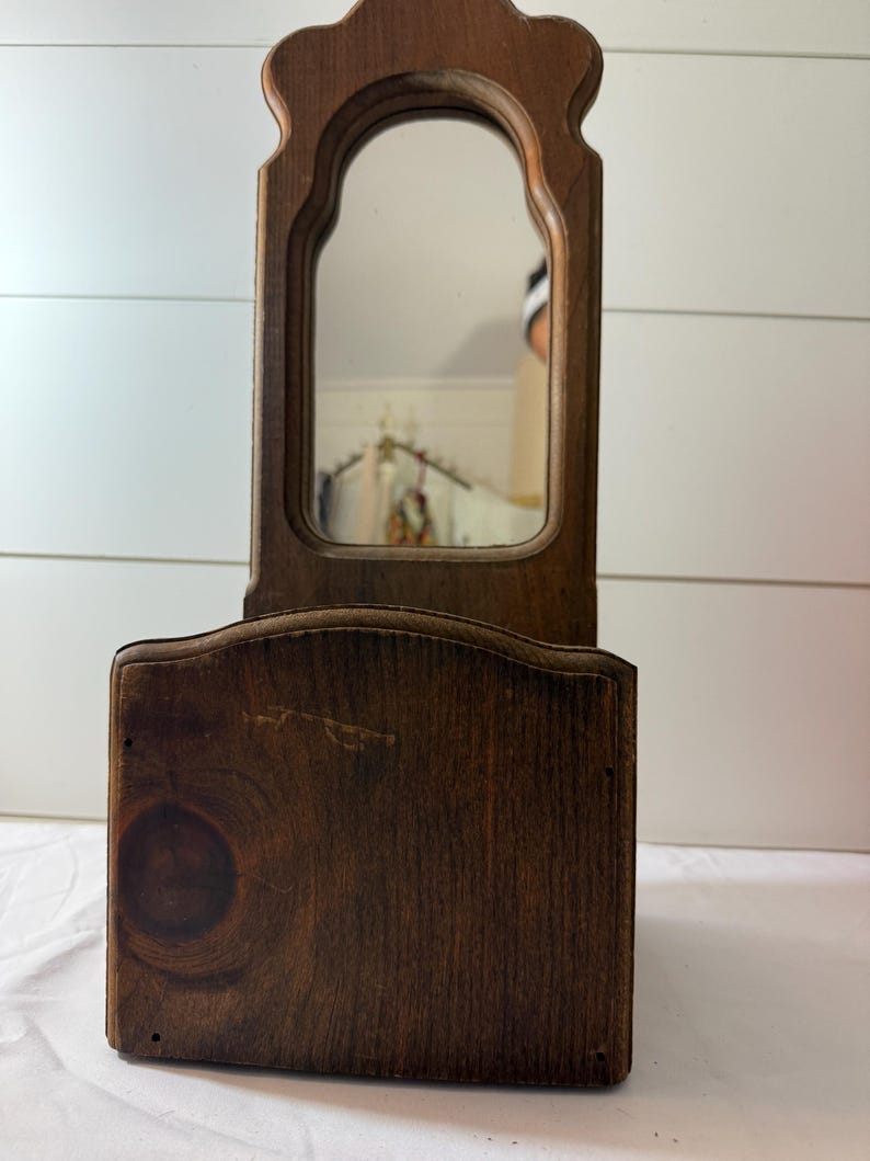 May include: A vintage wooden vanity mirror with a rectangular base and an arched mirror. The mirror is framed by a decorative wooden border. The wood has a rich, dark brown color and visible wood grain. May include: A vintage wooden vanity mirror with a rectangular base and an arched mirror. The mirror is framed by a decorative wooden border. The wood has a rich, dark brown color and visible wood grain.