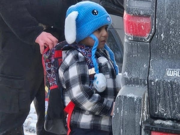 A young boy with a backpack and blue hat stands next to an SUV.