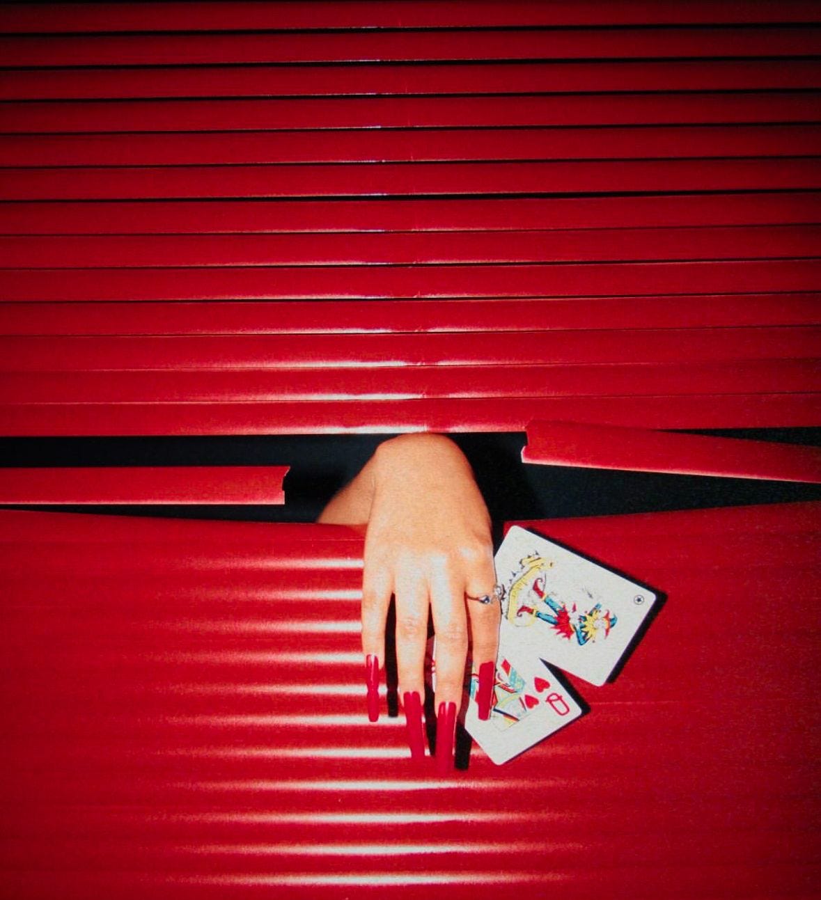 Hand with long red acrylic nails holding playing cards through red metal slats, bold red aesthetic fashion photography with high contrast lighting.