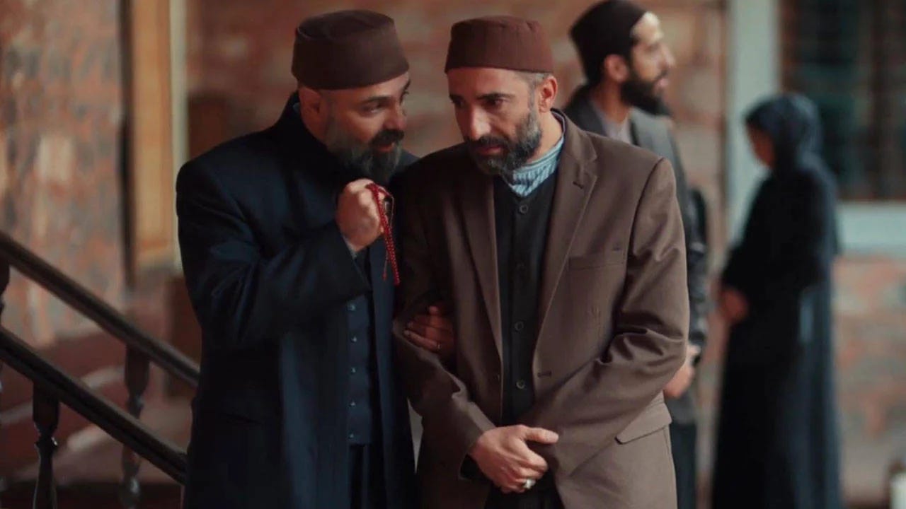 İki erkek karakter, tarihi bir mekanda konuşuyor ve biri elinde tesbih tutuyor. Arka planda geleneksel mimari detaylar ve diğer karakterler görülüyor.