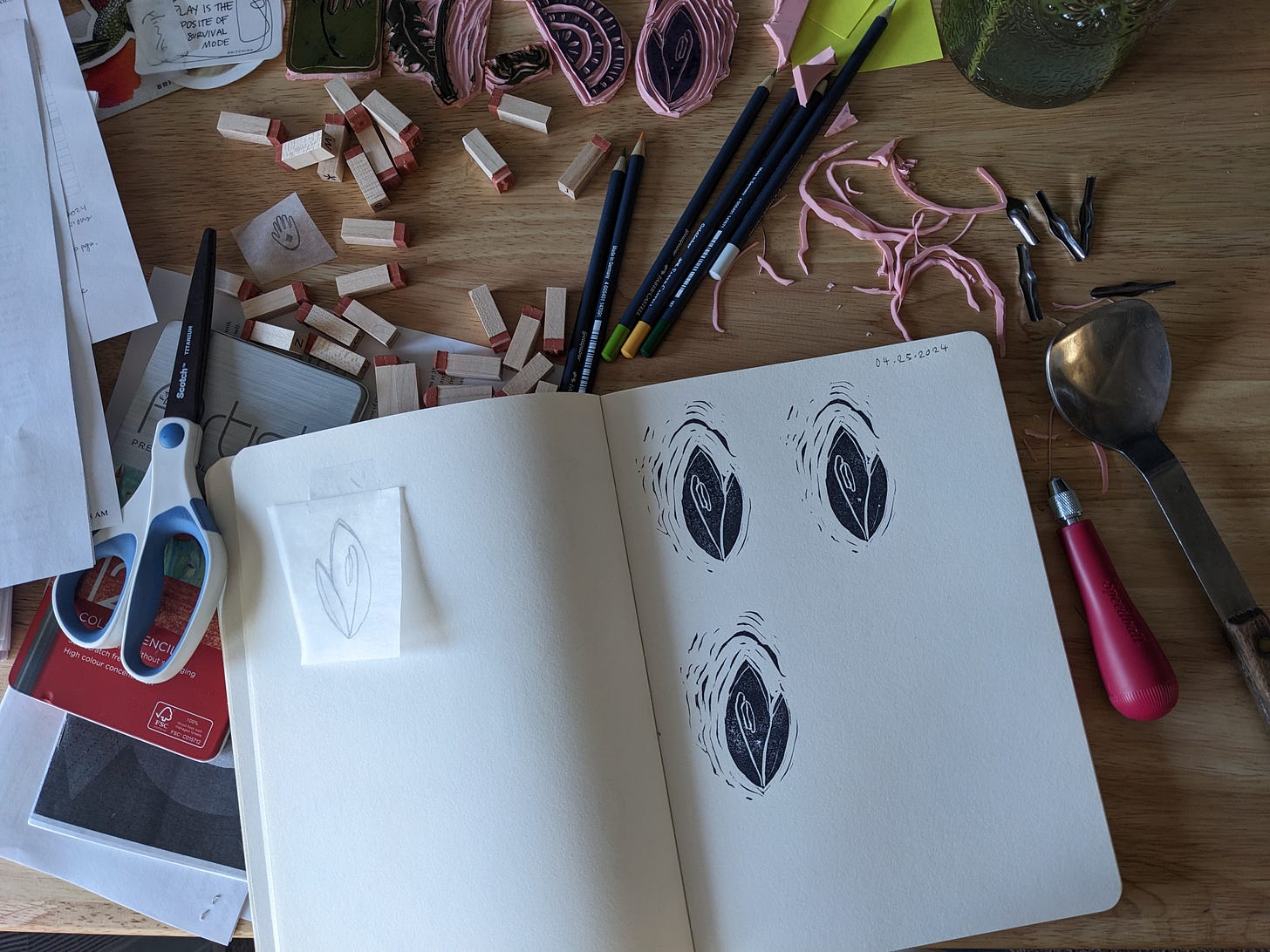 A photo of my messy desk, with stamps and scissors and block printing supplies spread out. A print of a trout lily is on an open sketchbook page. A photo of my messy desk, with stamps and scissors and block printing supplies spread out. A print of a trout lily is on an open sketchbook page.