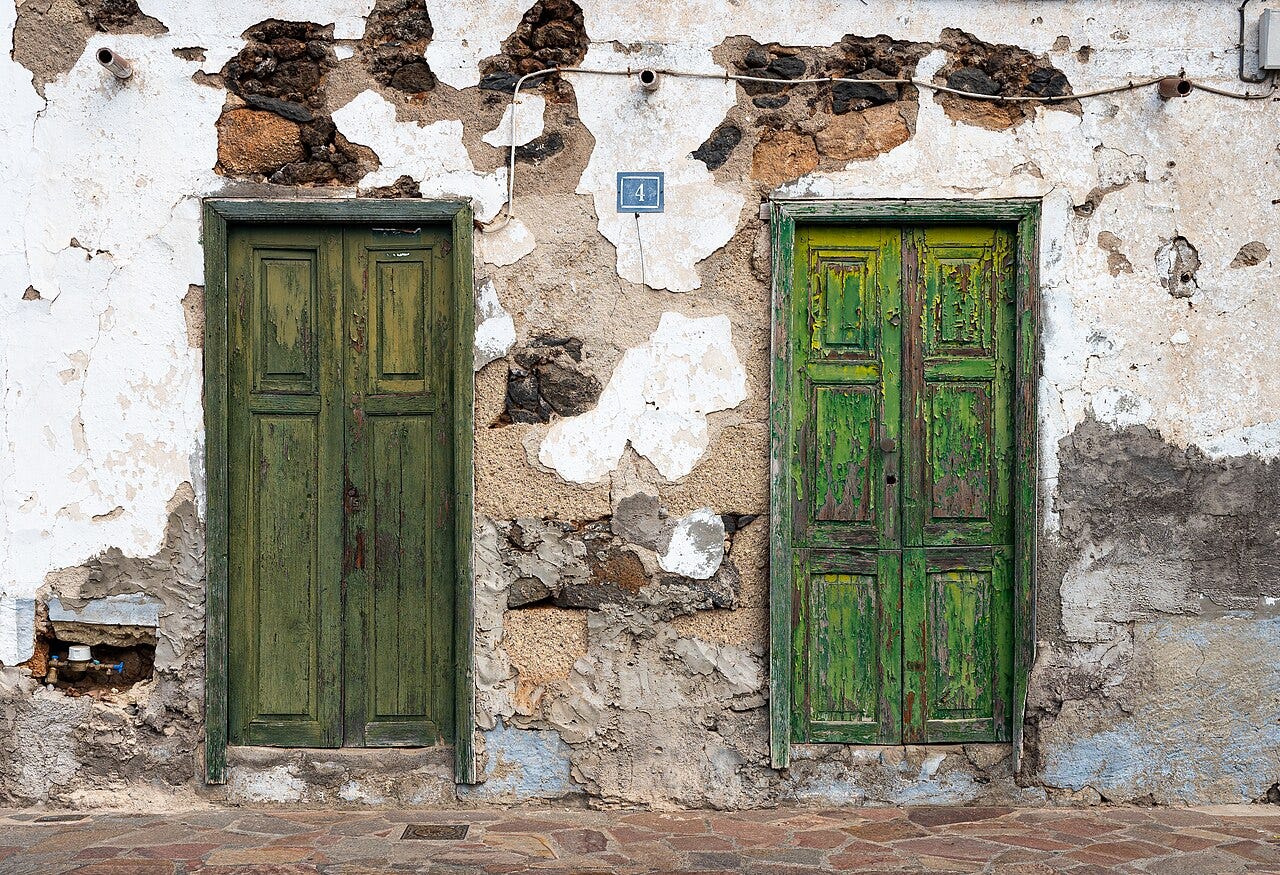 Two green doors in a wall that is decaying stucco.