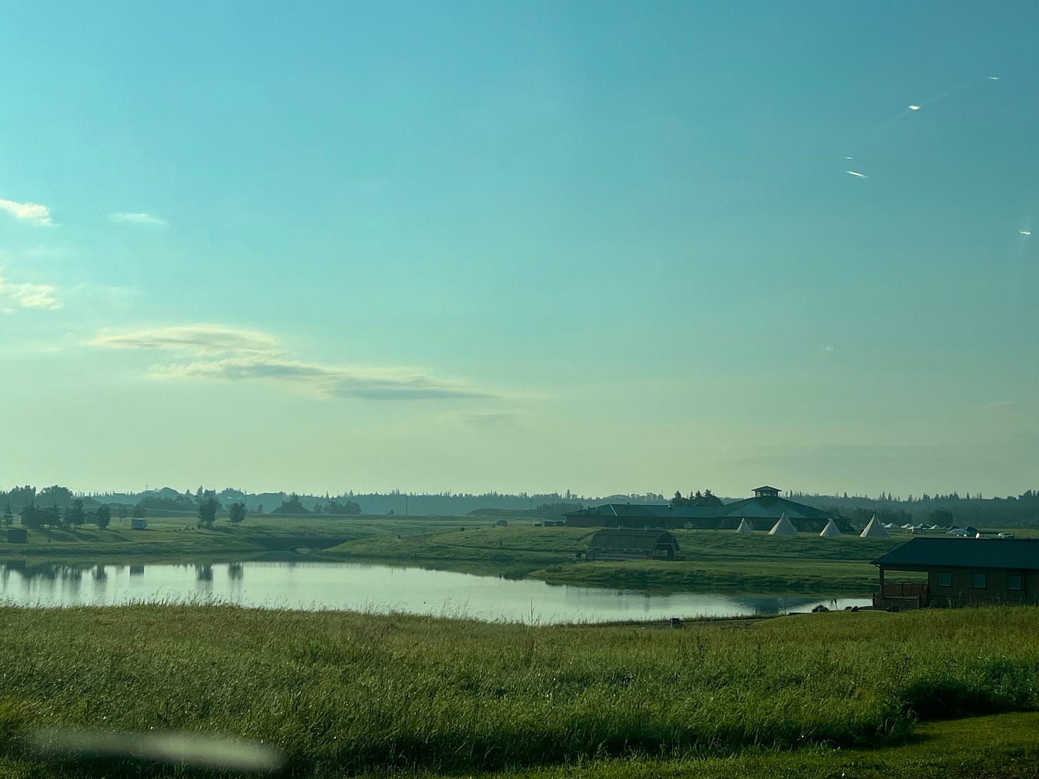 landscape photo with green grass in the foreground, a river in the middle and teepees and a lodge on the far side of the river. The sky is blue with a few clouds and the river is still, allowing reflections to show clearly.