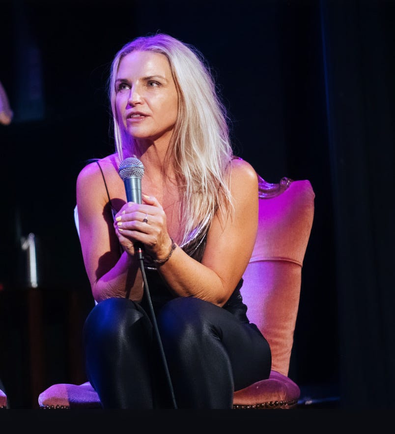 Jayne is sitting in an armchair onstage as a guest at a live event. She is holding a microphone
