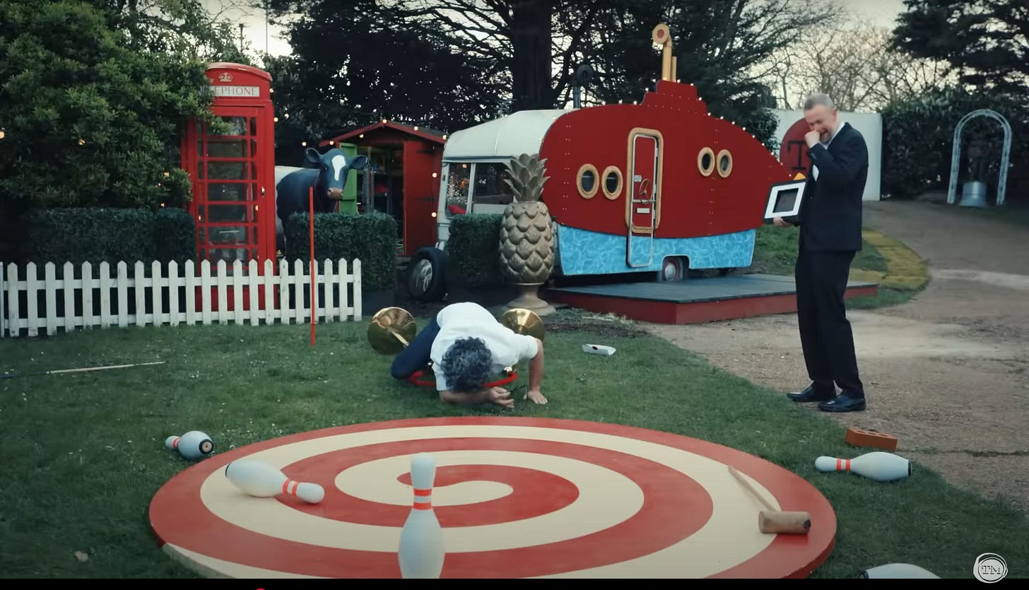 Jason Mantzoukas faceplants on the Taskmaster grounds while Little Alex Horne holds his fist to his mouth to suppress a giggle.