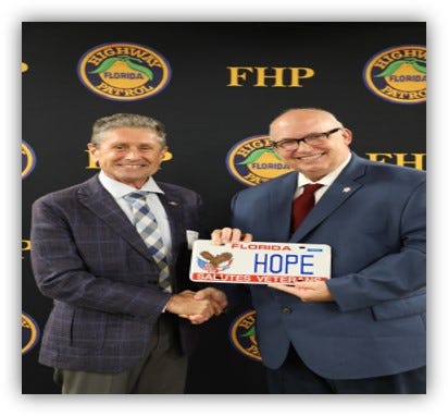 Two men in suits stand in front of a step‑and‑repeat backdrop featuring the Florida Highway Patrol (FHP) logo. They are shaking hands while holding a Florida specialty license plate that reads “FLORIDA HOPE – Salutes Veterans,” with an eagle and U.S. flag design. The scene appears to be an indoor ceremonial or promotional event.