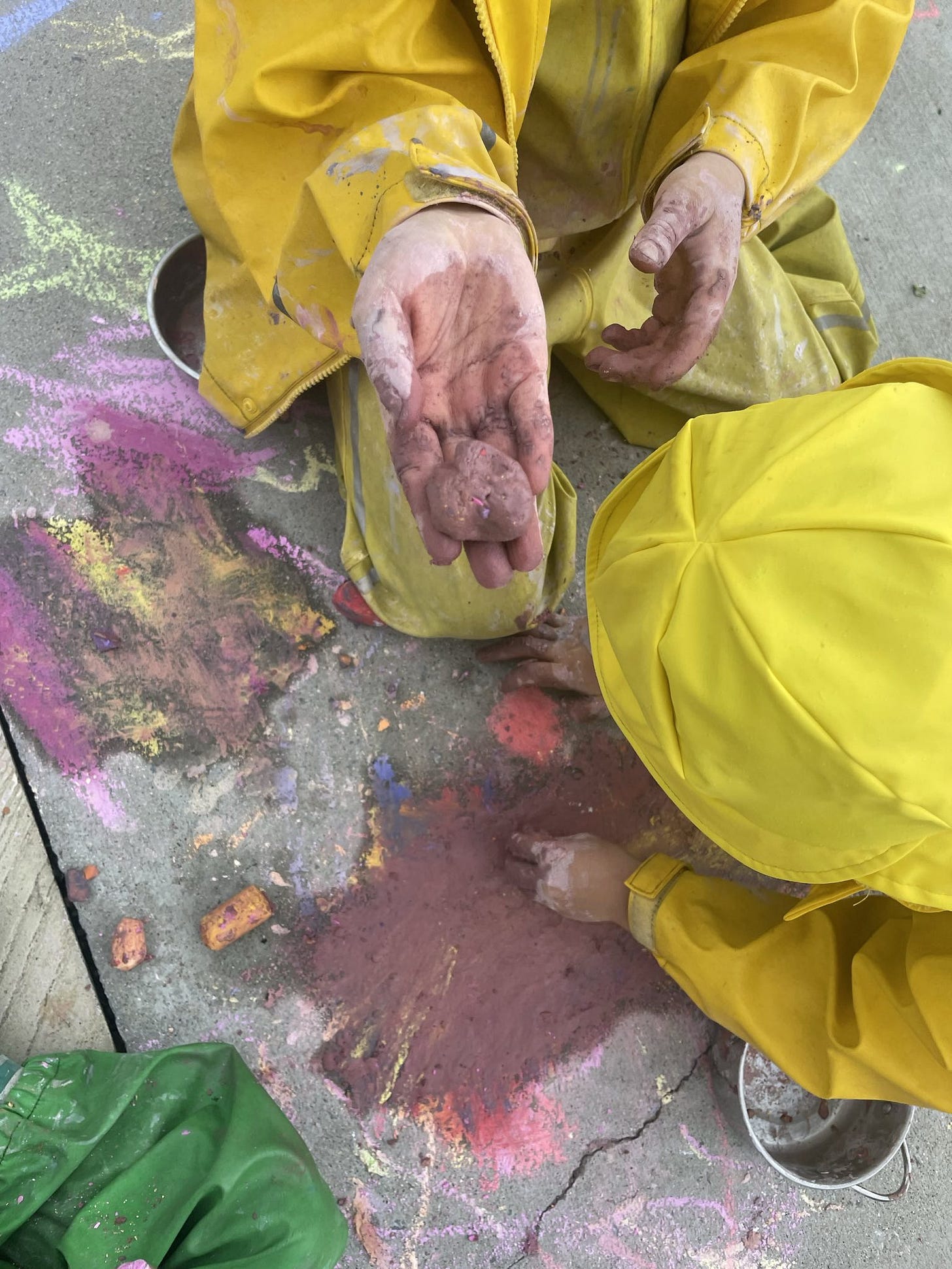 Two children wearing bright yellow rain gear are seated on the ground. They hold a block of chalk. On the ground around them are chalk drawings."