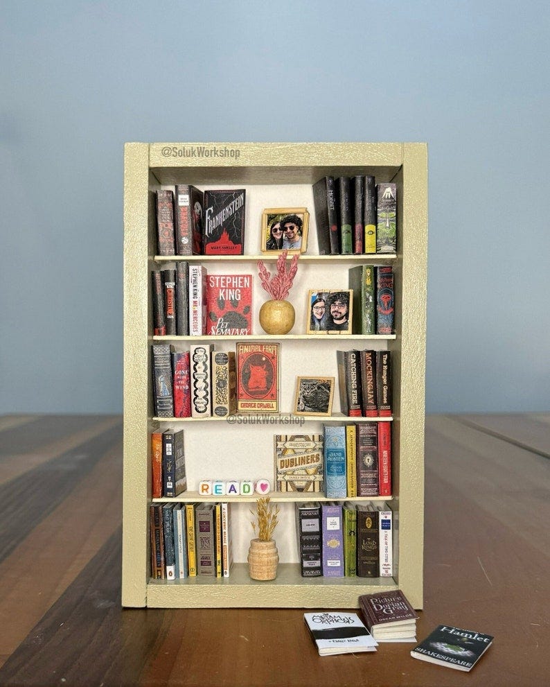 May include: A miniature bookshelf with a light brown wood frame and white shelves. The shelves are filled with miniature books with various titles, including 'Frankenstein', 'Stephen King', 'Pet Sematary', 'Finisher Fort', 'Dubliners', 'The Hunger Games', 'Catching Fire', 'Mockingjay', and 'Hamlet'. There are also miniature plants, a picture frame with a couple, and a heart-shaped object on the shelves. The bookshelf is sitting on a wooden surface.