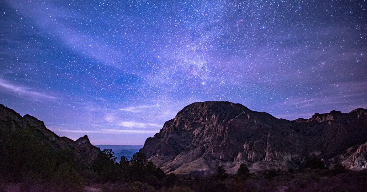  A clear night sky filled with stars above a rugged desert canyon, with rocky mountain silhouettes under soft purple and blue hues.